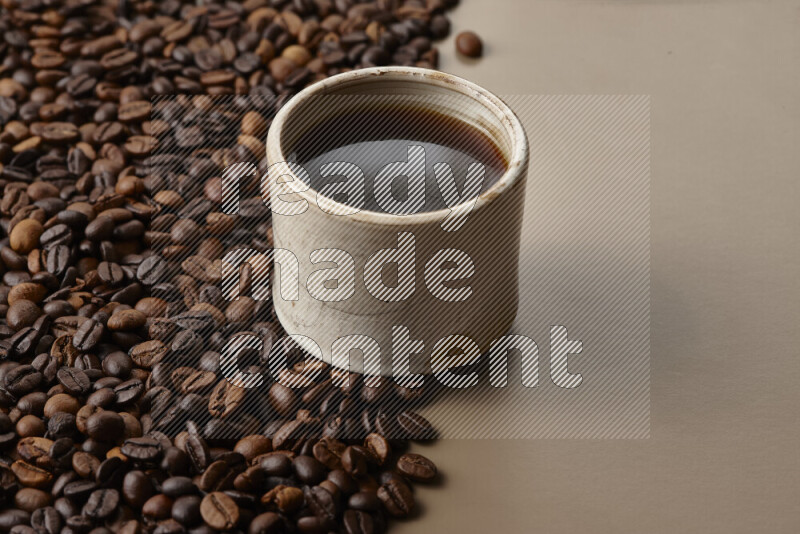 A beige pottery cup of coffee surrounded by roasted coffee beans on beige background