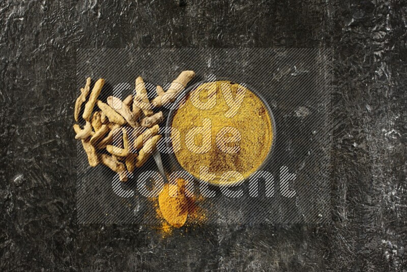A pottery black bowl and a metal spoon full of turmeric powder with dried turmeric fingers on a textured black flooring