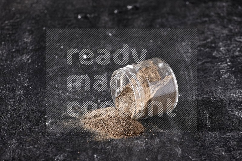 A flipped glass jar full of black pepper powder on a textured black flooring