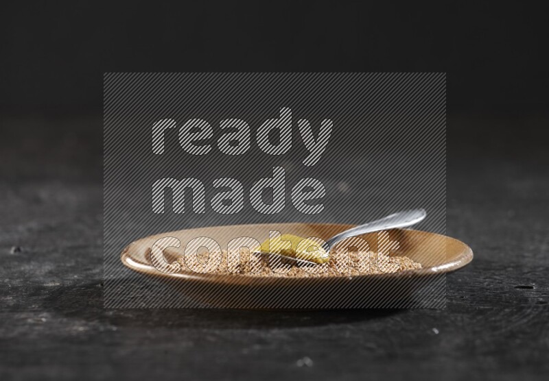 صحن فخار بيج ممتلئ ببذور الخردل مع ملعقة معدنية ممتلئة بمعجون الخردل داخل الصحن على خلفية سوداء