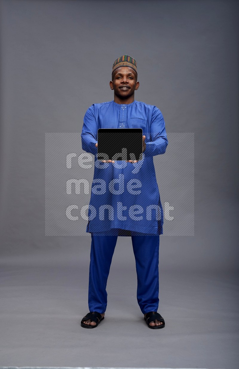 Man wearing Nigerian outfit standing showing tablet to camera on gray background