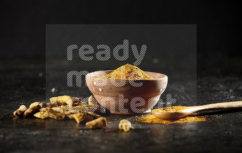 A wooden bowl and a wooden spoon full of turmeric powder with dried turmeric fingers on textured black flooring