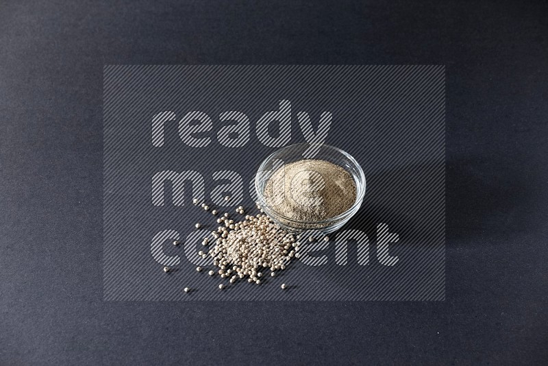 A glass bowl full of white pepper powder with white pepper beads on black flooring