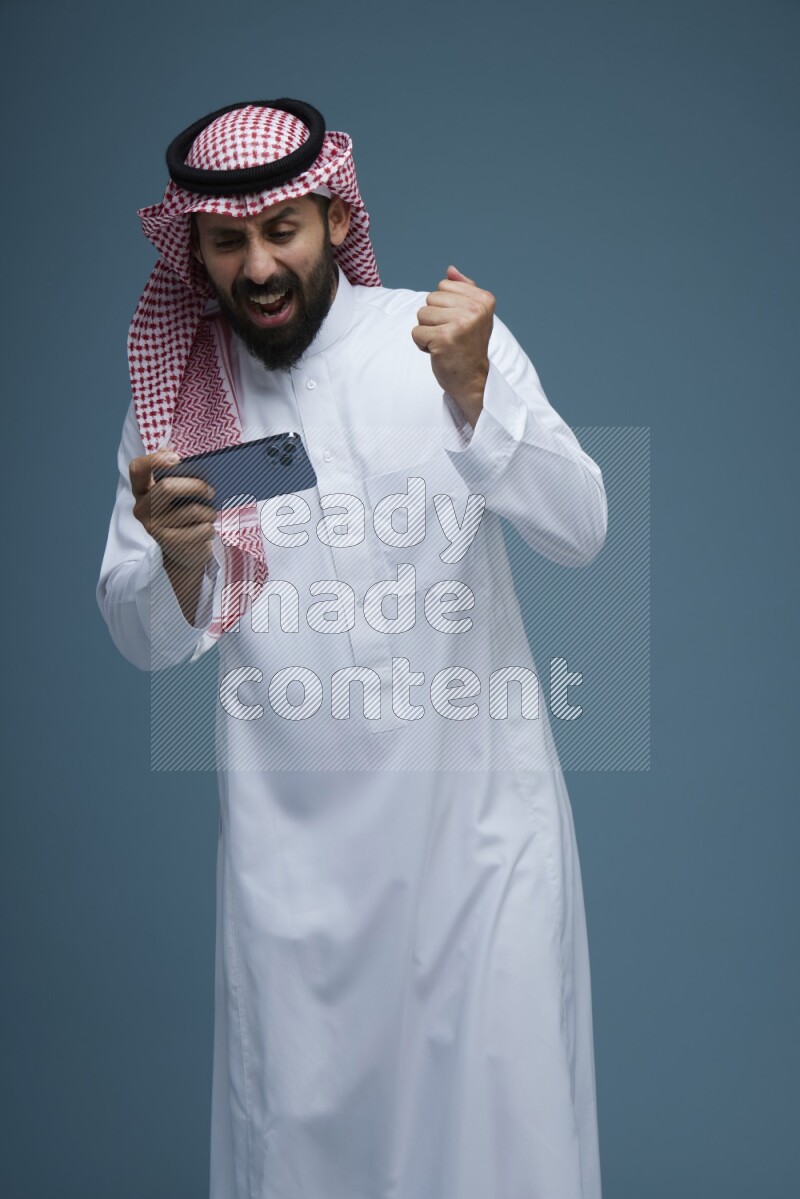A man Playing a Game on his phone in a blue background wearing Saudi Thob with Shomag