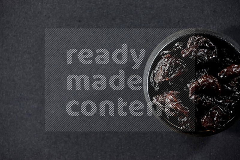 A black pottery bowl full of dried plums on a black background in different angles
