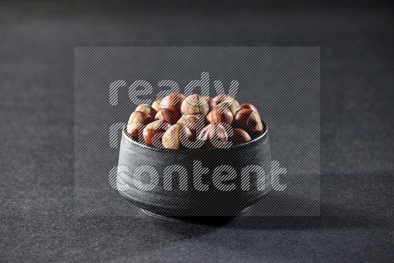 A black pottery bowl full of hazelnuts on a black background in different angles