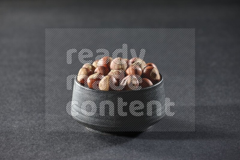 A black pottery bowl full of hazelnuts on a black background in different angles