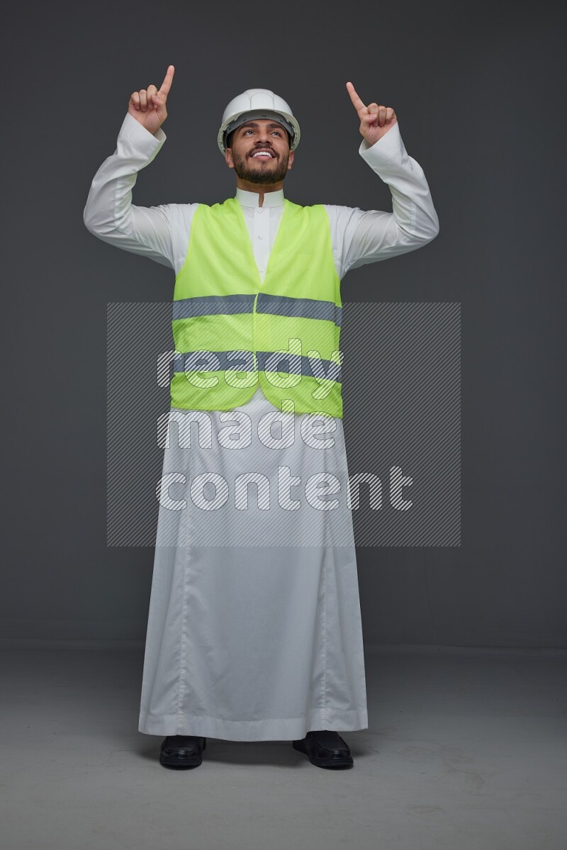 A Saudi man wearing Thobe with a yellow safety vest and white helmet standing and pointing different angles eye level on a gray background