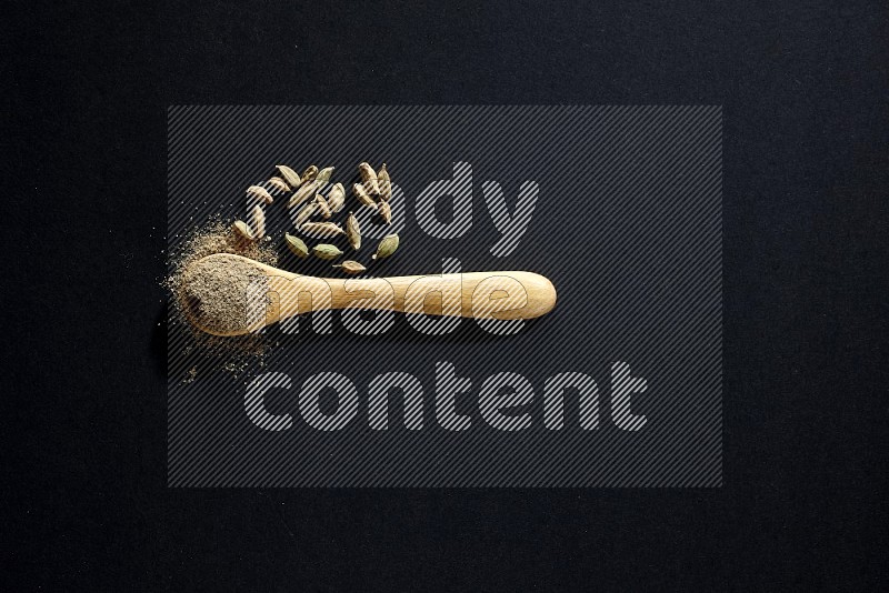 ملعقة خشبية ممتلئة ببودرة الهيل وحبوب الهيل بجانبها على خلفية سوداء