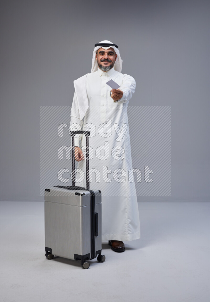 Saudi man wearing Thob and white Shomag standing holding Travel bag and ATM card on Gray background