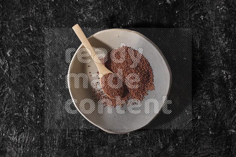 A multicolored pottery plate full of garden cress seeds and wooden spoon full of seeds on a textured black flooring