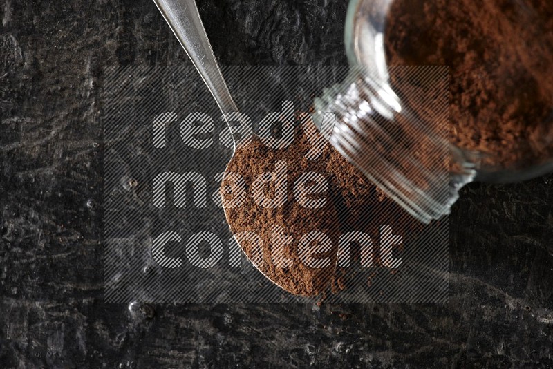 A flipped glass spice jar and a metal spoon full of cloves powder on textured black flooring