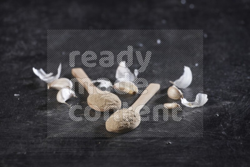 Two wooden spoons filled with garlic powder, surrounded by peeled garlic cloves and their skins on a textured black flooring