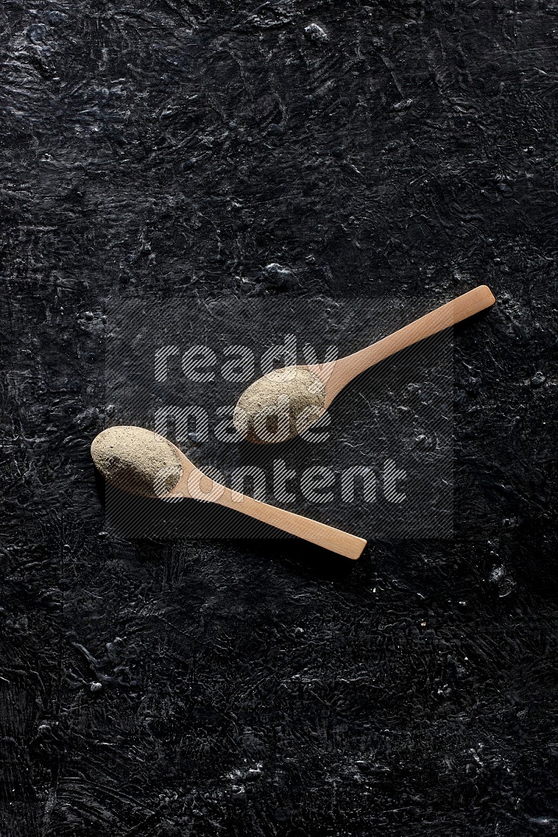 2 wooden spoons full of white pepper powder on textured black flooring