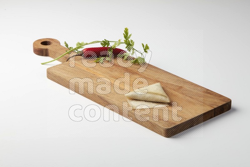 One uncooked samosa with a single red pepper and parsley on a wooden cutter on a white background