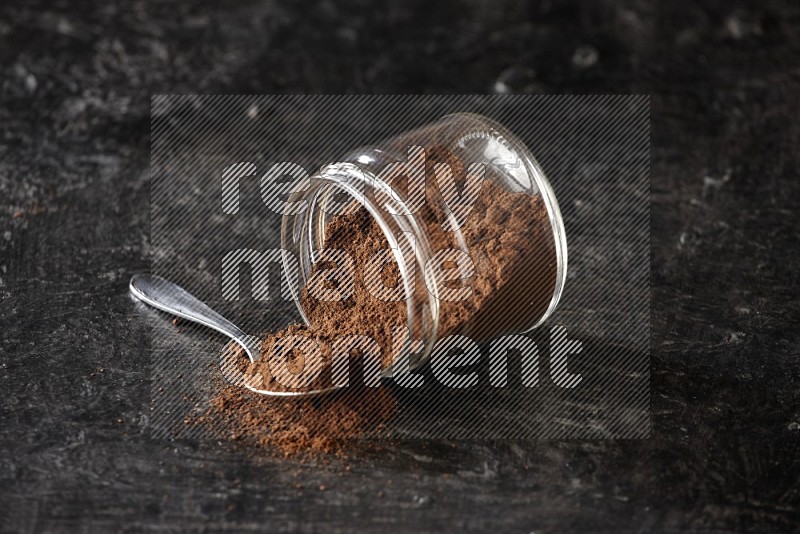 A flipped glass jar and metal spoon full of cloves powder with cloves spread on a textured black flooring