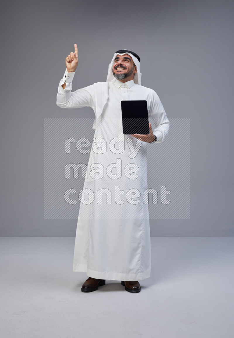 Saudi man Wearing Thob and white Shomag standing showing tablet to camera on Gray background