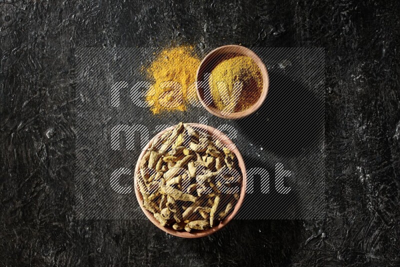 2 wooden bowls, one full of turmeric powder and the other full of dried turmeric whole fingers on a textured black flooring