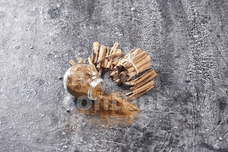 Herbal glass jar full of cinnamon powder flipped with cinnamon sticks stacked and bounded on a textured black background