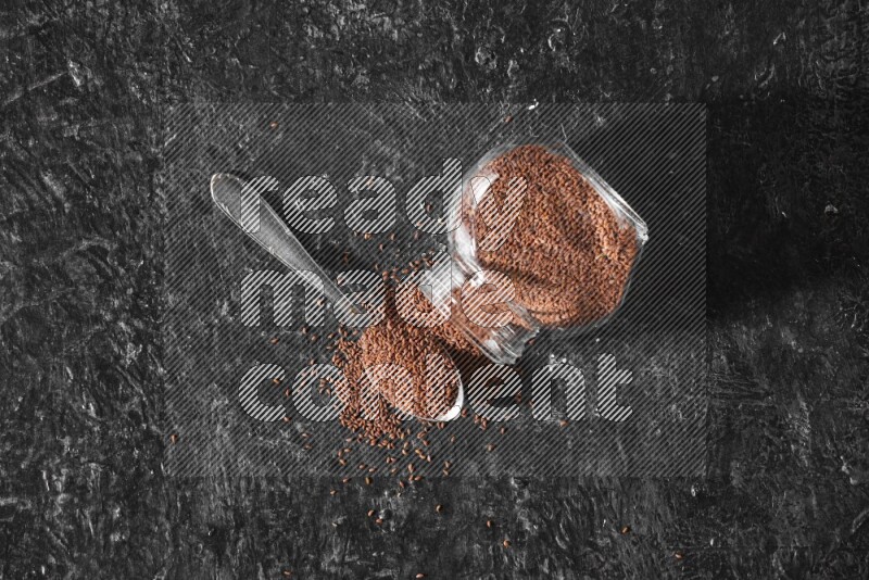 A glass spice jar full of garden cress seeds flipped and seeds spread out with a metal spoon full of the seeds on a textured black flooring
