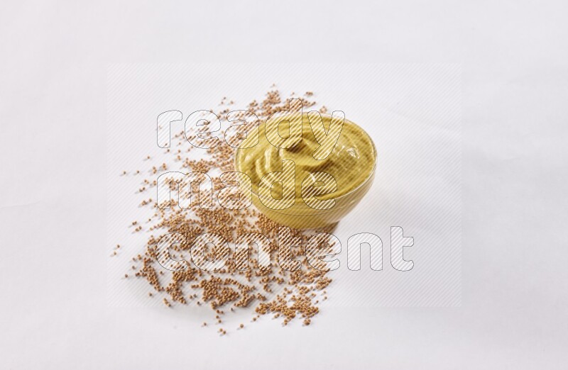 A glass bowl full of mustard paste with mustard seeds underneath on white flooring
