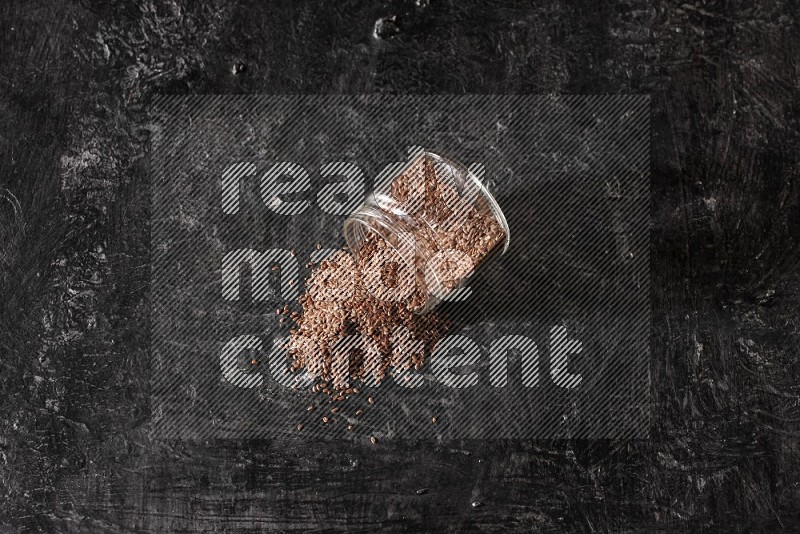 A glass jar full of flaxseeds flipped and seeds spread out on a textured black flooring