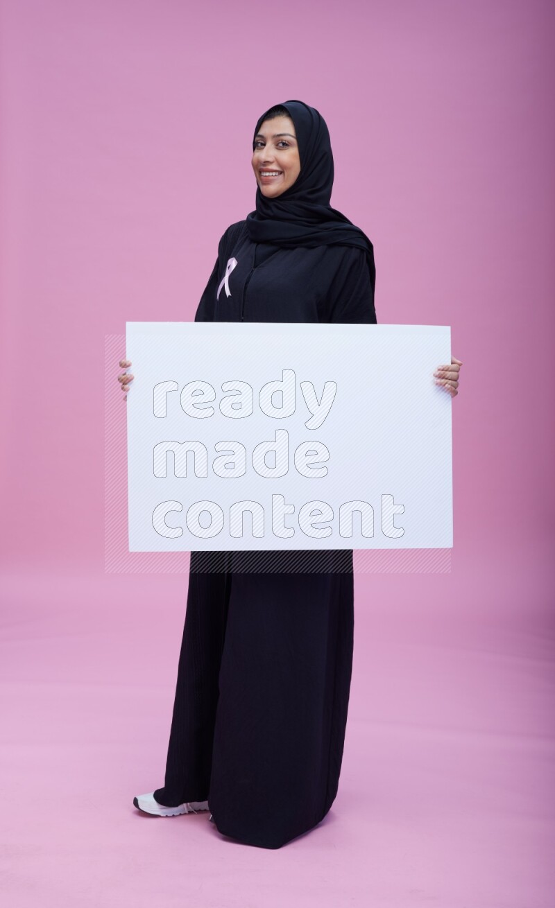 Saudi woman wearing pink ribbon on Abaya standing holding board on pink background