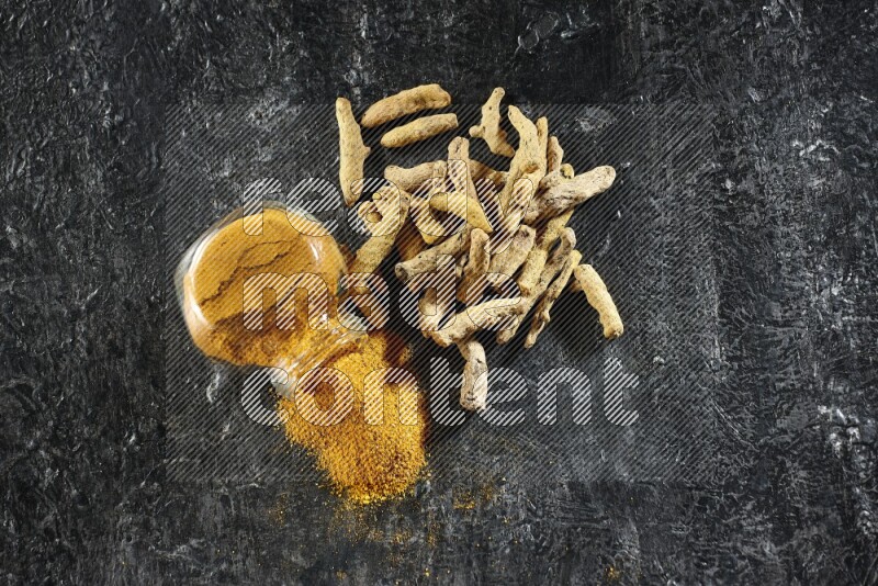 A flipped glass spice jar full of turmeric powder and powder spilled out of it with dried whole fingers on textured black flooring