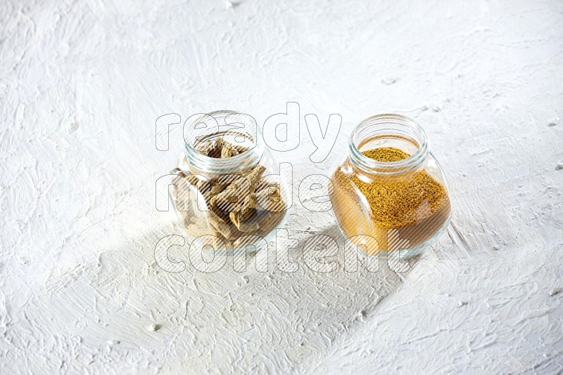 2 glass spice jars full of turmeric powder and dried whole fingers on textured white flooring
