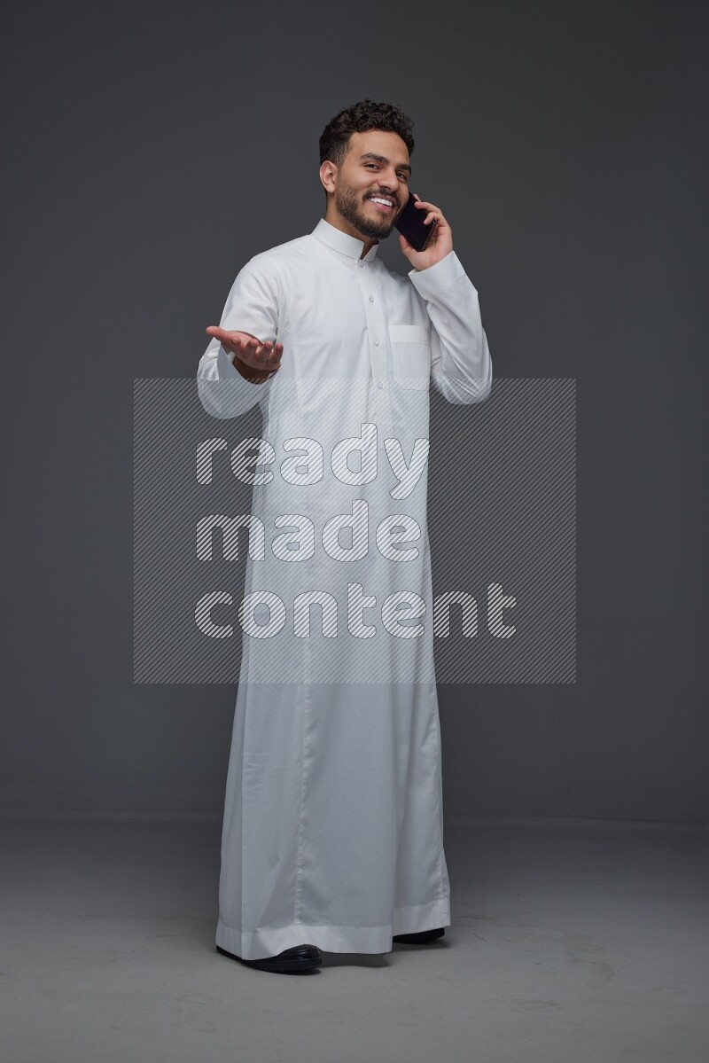 A Saudi man wearing Thobe and talking in the phone while standing and making different poses eye level on a gray background
