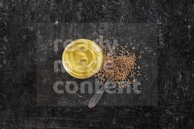 A glass jar full of mustard paste and a metal spoon full of mustard seeds on a textured black flooring