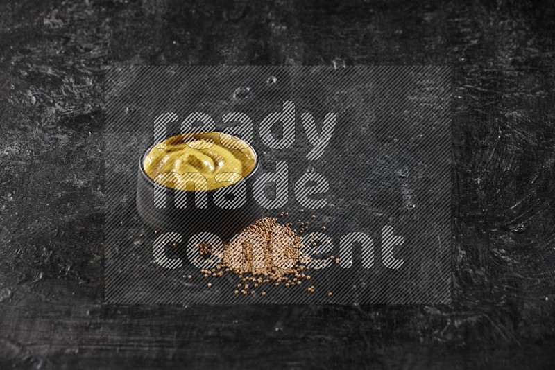 A black pottery bowl full of mustard paste with mustard seeds underneath on textured black flooring in different angles