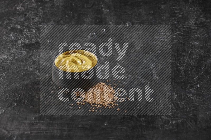 A black pottery bowl full of mustard paste with mustard seeds underneath on textured black flooring