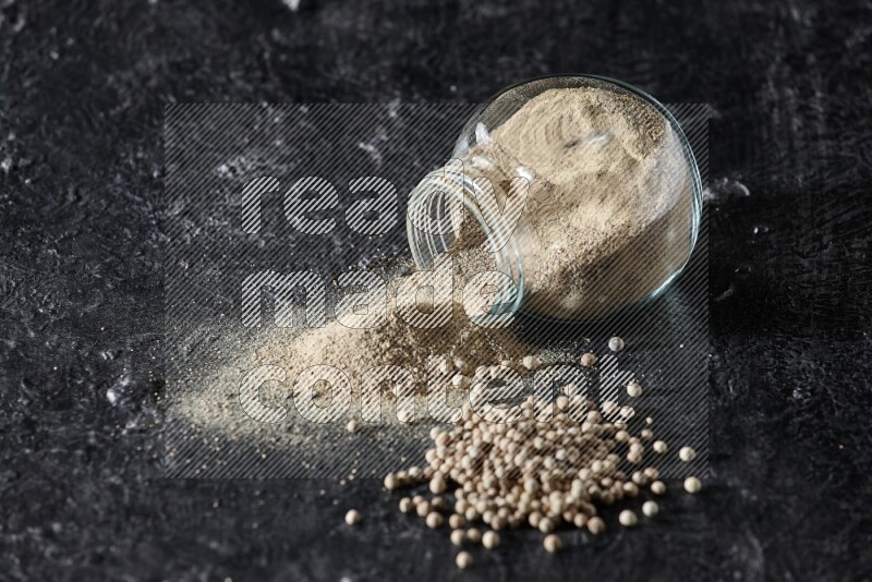 A flipped herbal glass jar full of white pepper powder with spilled powder and pepper beads on textured black flooring
