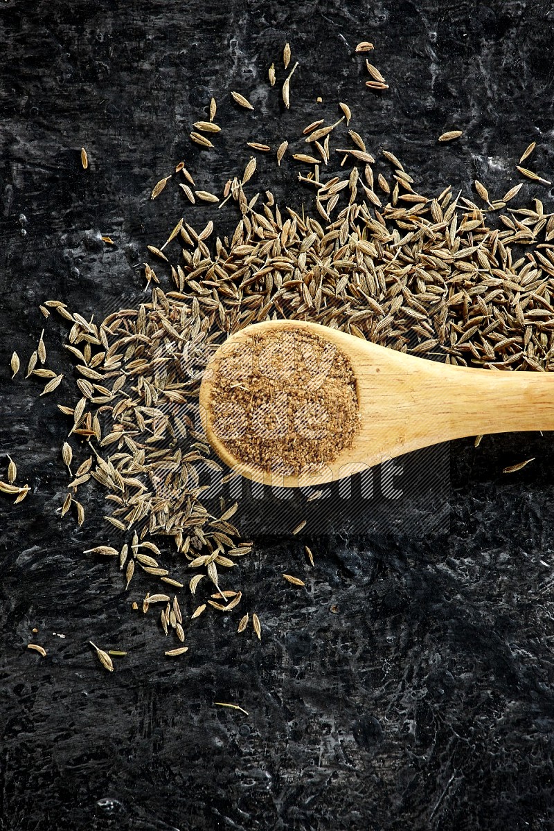 A wooden spoon full of cumin powder and cumin seeds spreaded on textured black flooring
