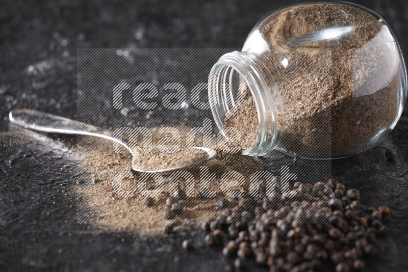 A flipped glass spice jar full of black pepper powder with a metal spoon full of powder and pepper beads on textured black flooring
