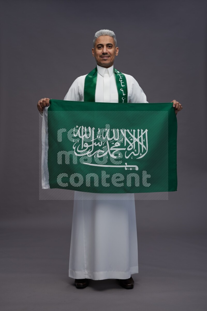 Saudi man wearing thob standing holding Saudi flag on gray background