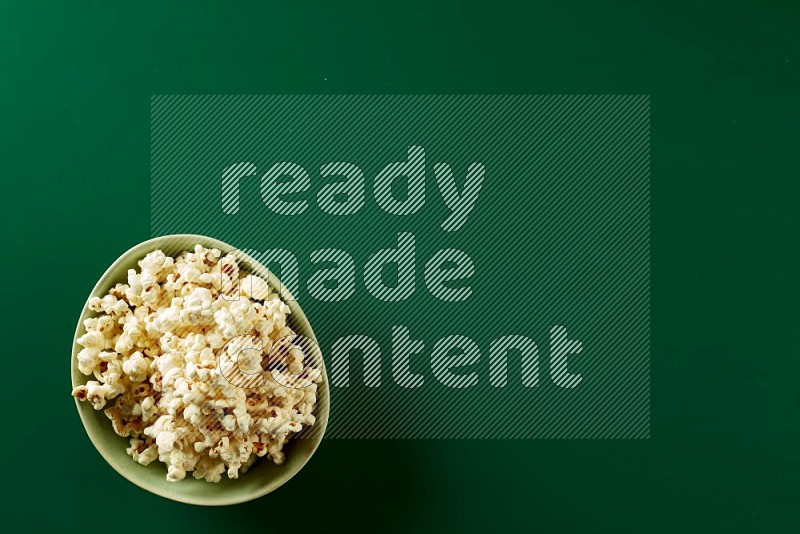 A green ceramic bowl full of popcorn on a green background in a top view shot