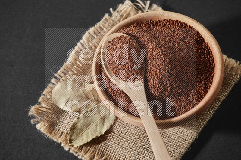 A wooden bowl full of garden cress seeds with wooden spoon full of the seeds on it on burlap fabric on a black flooring