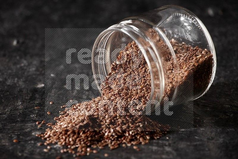 A glass jar full of flaxseeds flipped and seeds spread out on a textured black flooring