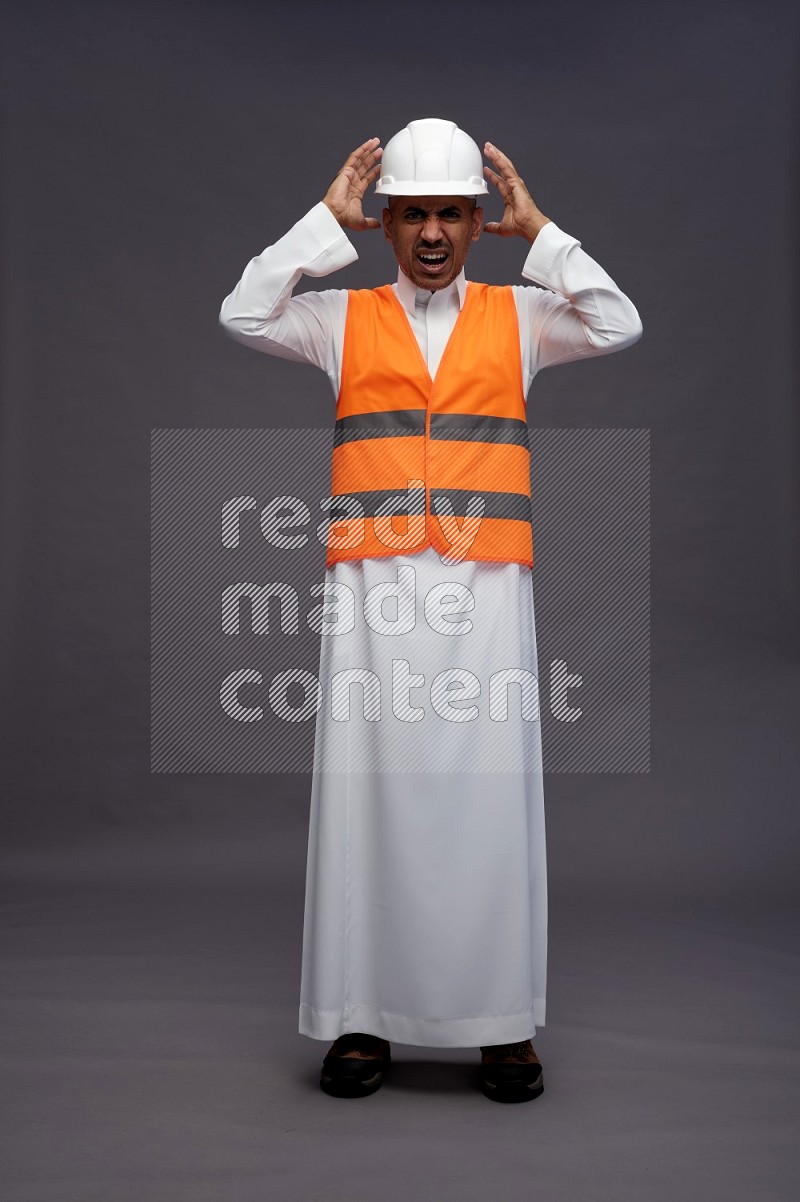 Saudi man wearing thob with engineer vest standing hands on head on gray background