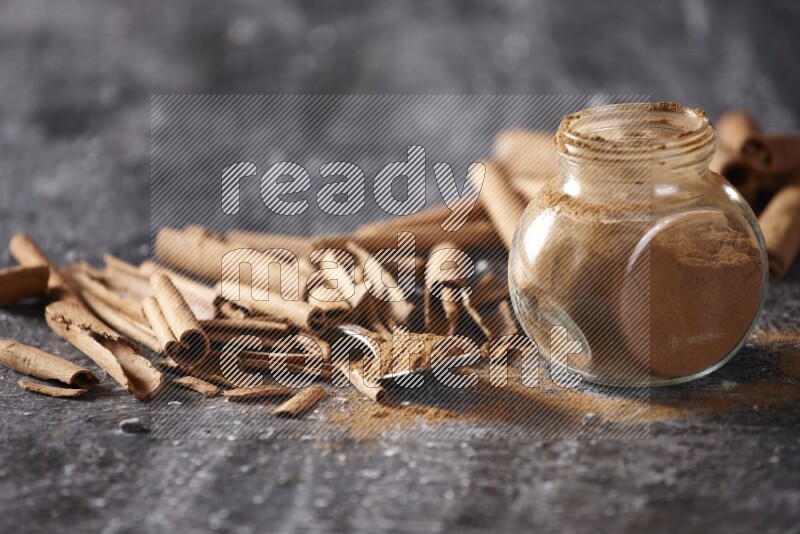 Herbal glass jar and a metal spoon full of cinnamon powder surrounded by cinnamon sticks on textured black background
