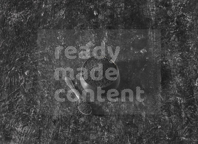 A black pottery bowl full of black seeds with a metal spoon full of the seeds on a textured black flooring