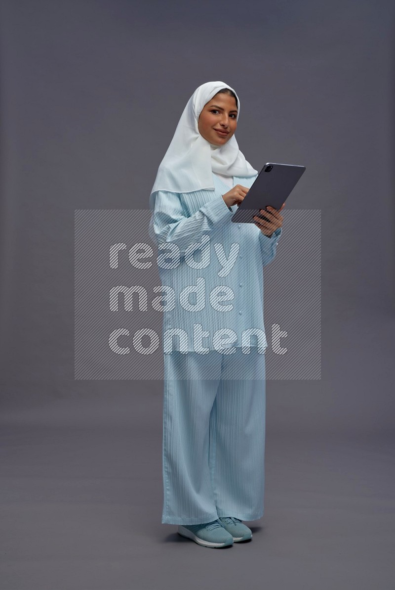 Saudi woman wearing hijab clothes standing working on tablet on gray background