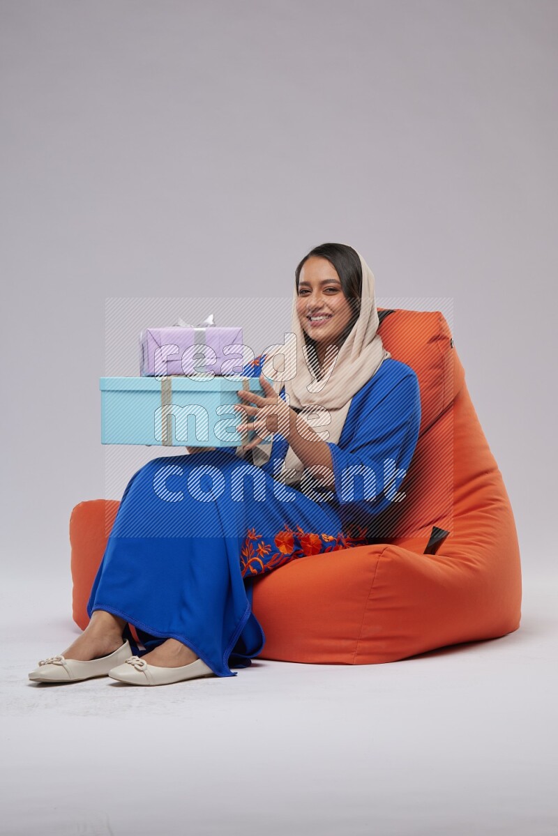 A woman sitting on an orange beanbag wearing Jalabeya holding a gift box