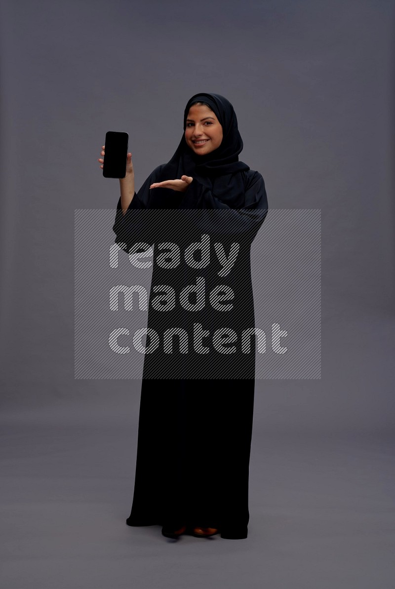 Saudi woman wearing Abaya standing showing phone to camera on gray background