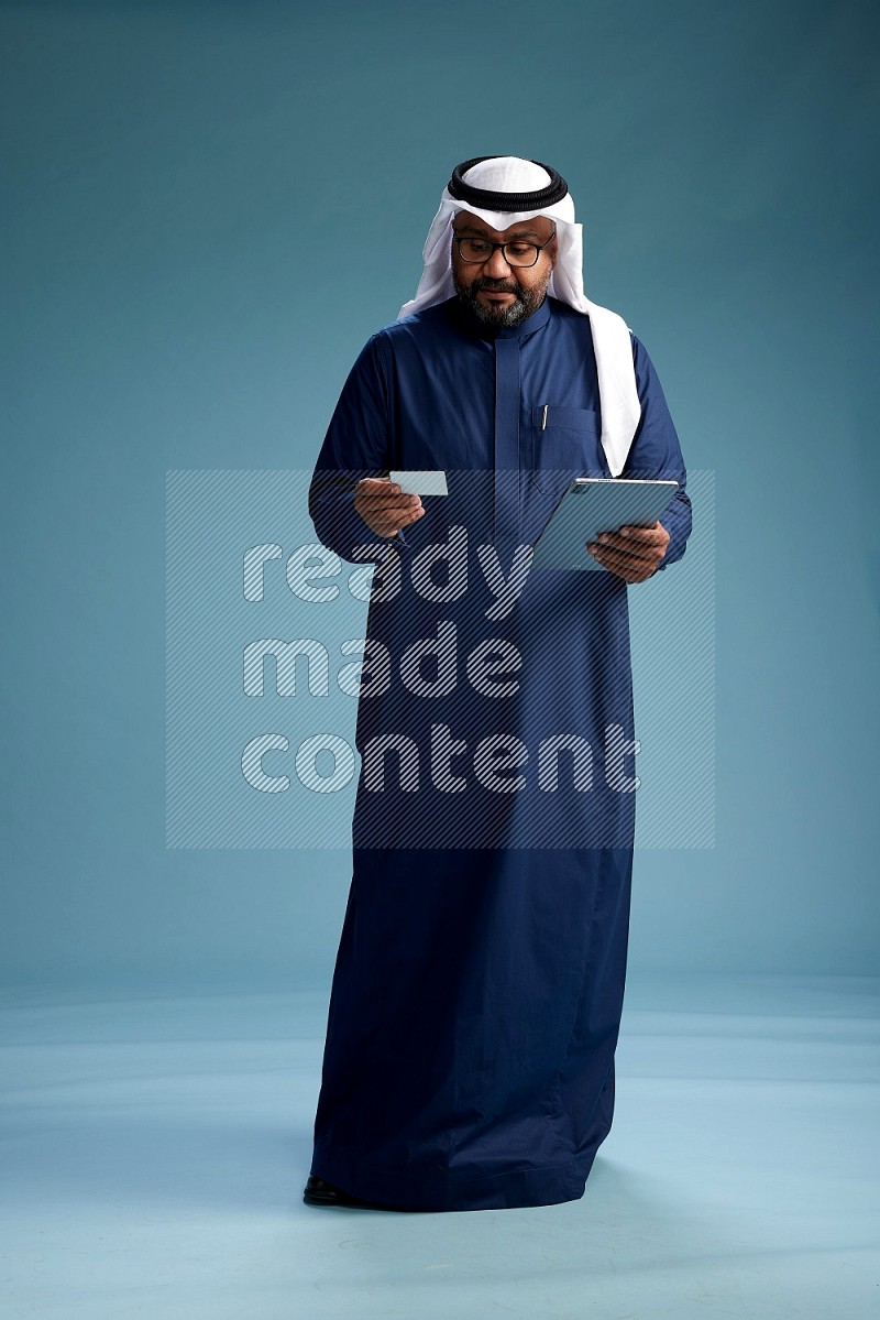 Saudi Man with shimag Standing holding ATM card while working on tablet on blue background