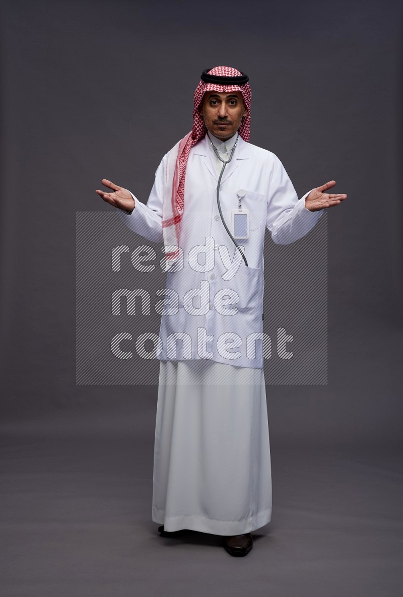 Saudi man wearing thob with lab coat and shomag with pocket employee badge with stethoscope standing interacting with the camera on gray background