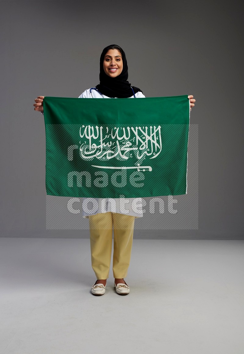Saudi woman wearing lab coat with stethoscope standing holding Saudi flag on Gray background