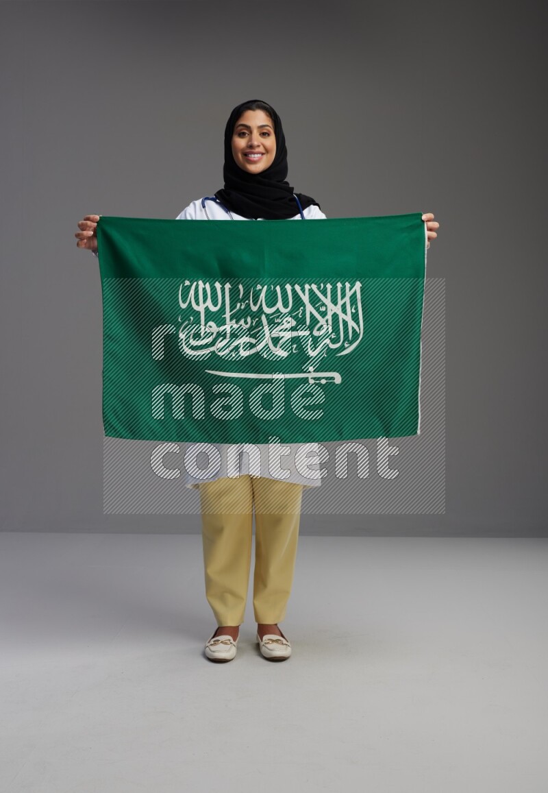 Saudi woman wearing lab coat with stethoscope standing holding Saudi flag on Gray background